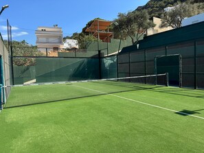 A tennis court with a net, surrounded by a green fence and a building in the background.