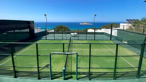 Un court de tennis avec un filet, entouré d’une clôture verte, offrant une vue sur la côte, avec la mer et une petite île au loin.