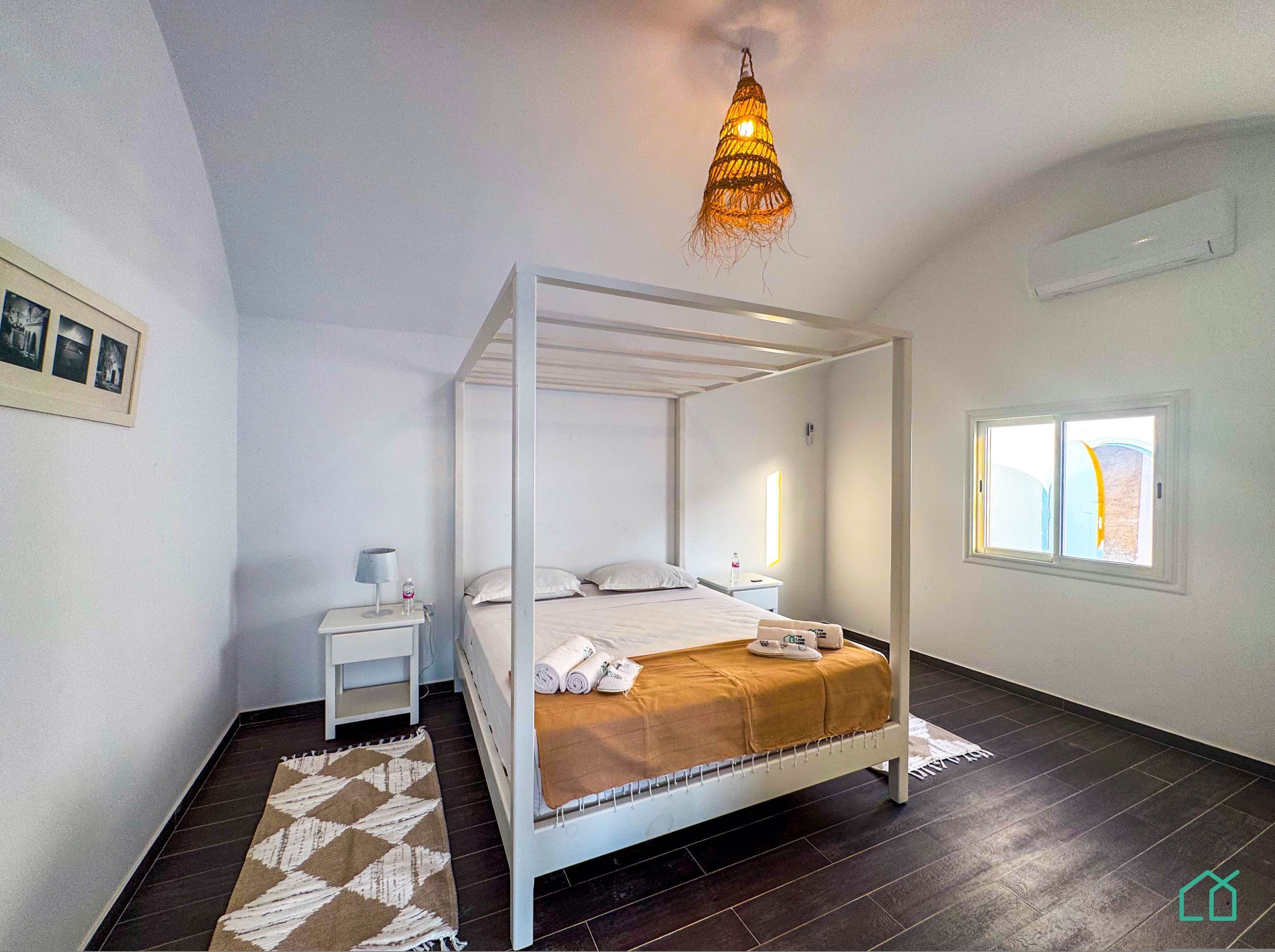 Une chambre moderne avec un lit à baldaquin, une table de chevet avec une lampe, une petite commode, une fenêtre donnant sur un beau paysage et un tapis à motifs sur le plancher en bois foncé.