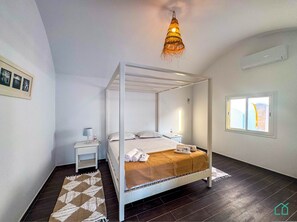A modern bedroom with a four-poster bed, a bedside table with a lamp, a small nightstand, a window with a view, and a patterned rug on the dark wooden floor. - Padel Resort 2bd Apt (Raf Raf)