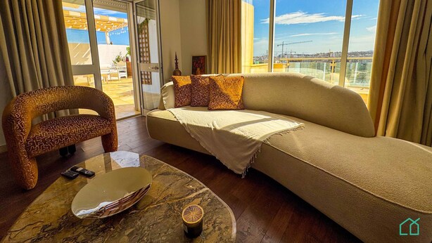 Una sala de estar moderna con un sofá, un sillón estampado, una mesa de centro de mármol y vistas de la ciudad a través de grandes ventanales.