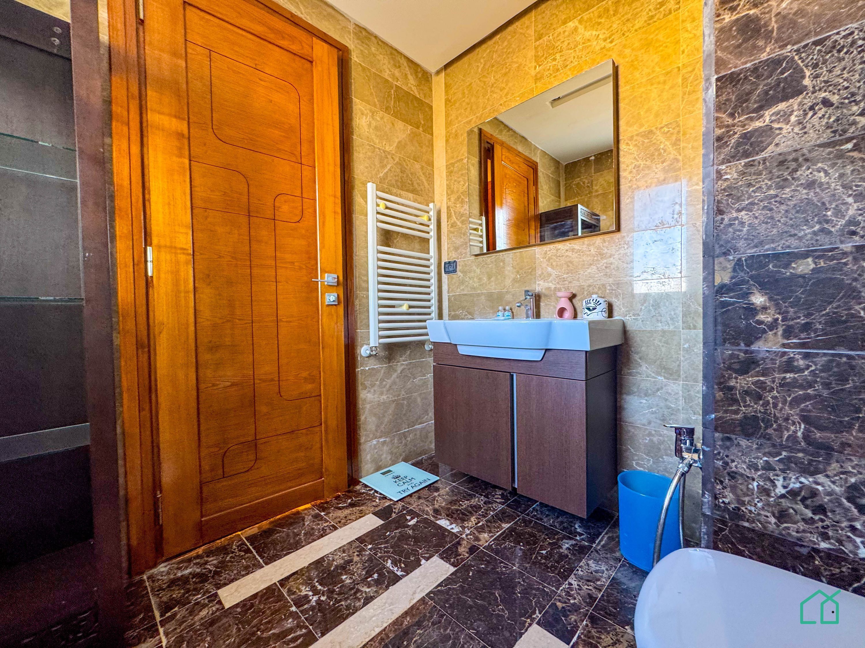 A bathroom with a wooden door, a white sink, a mirror, and a blue trash bin.