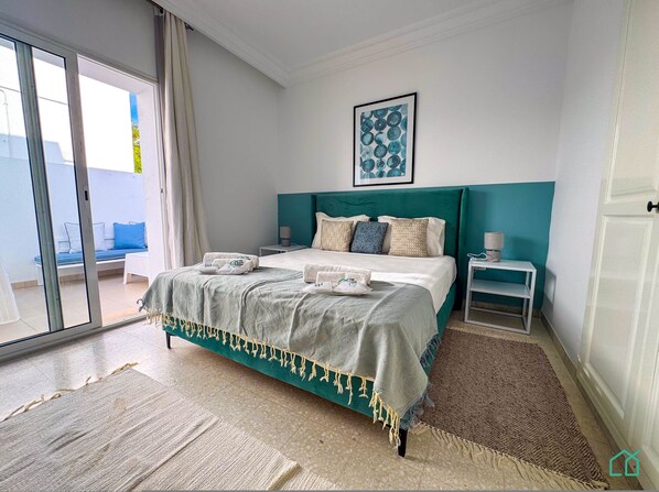 A bedroom with a bed, a nightstand, a window with curtains, a wall-mounted artwork, and a view of the outside. - Coste Sidi Bou Said A2 Beautiful and Central (Sidi Bou Said)