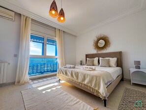 Un dormitorio con una cama grande, una ventana con mosquitera azul, un aplique de techo y un espejo fijado a la pared.