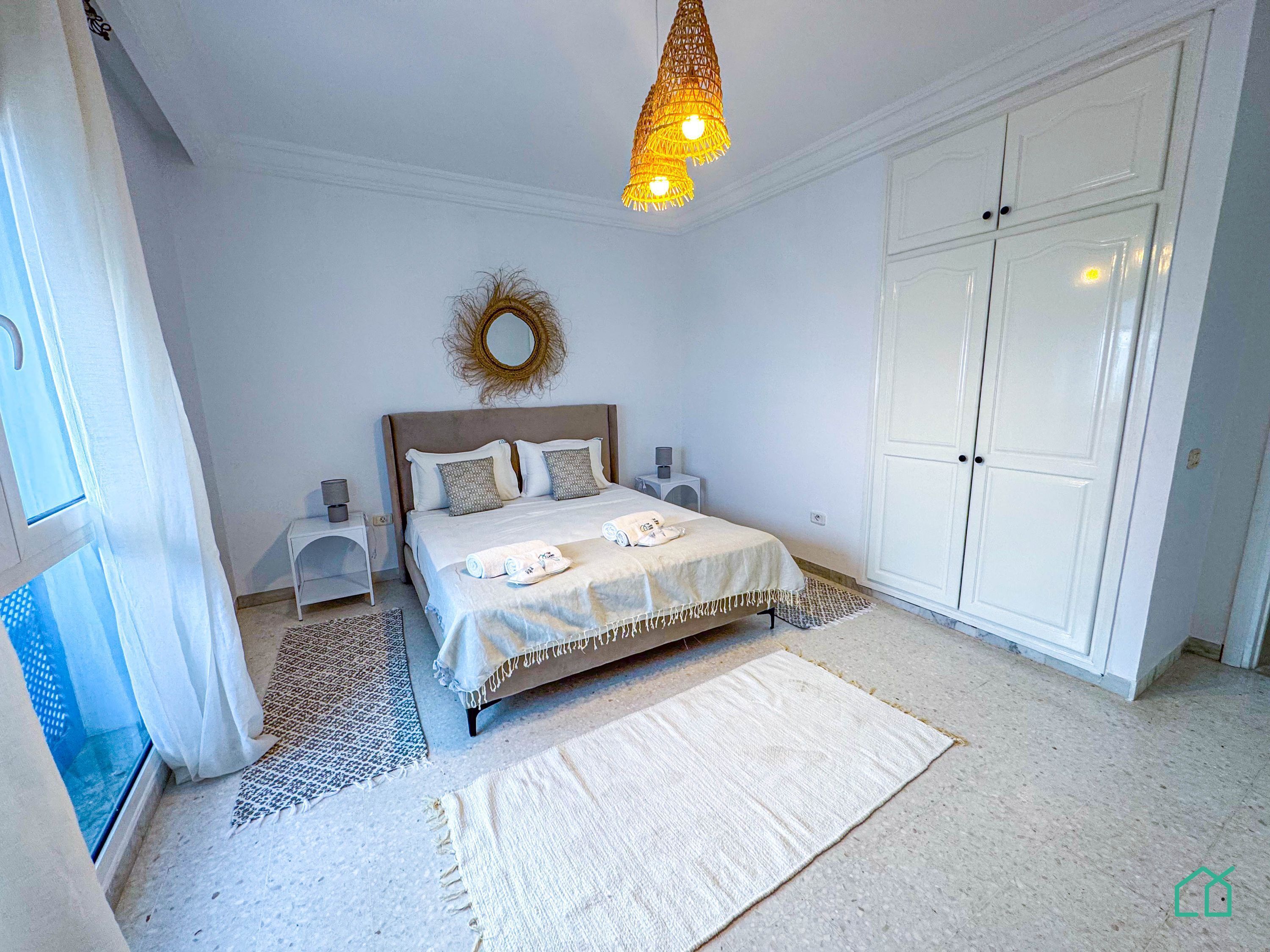 A bedroom with a bed, bedside tables, a hanging light fixture, a wardrobe, and a mirror on the wall.