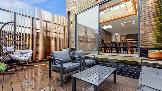 A wooden deck with outdoor seating, including a swing chair and sofas, and a view into a modern interior space with a dining area. - Luxury Abode: 5-Bed Haven Ideal for Big Groups! (London)