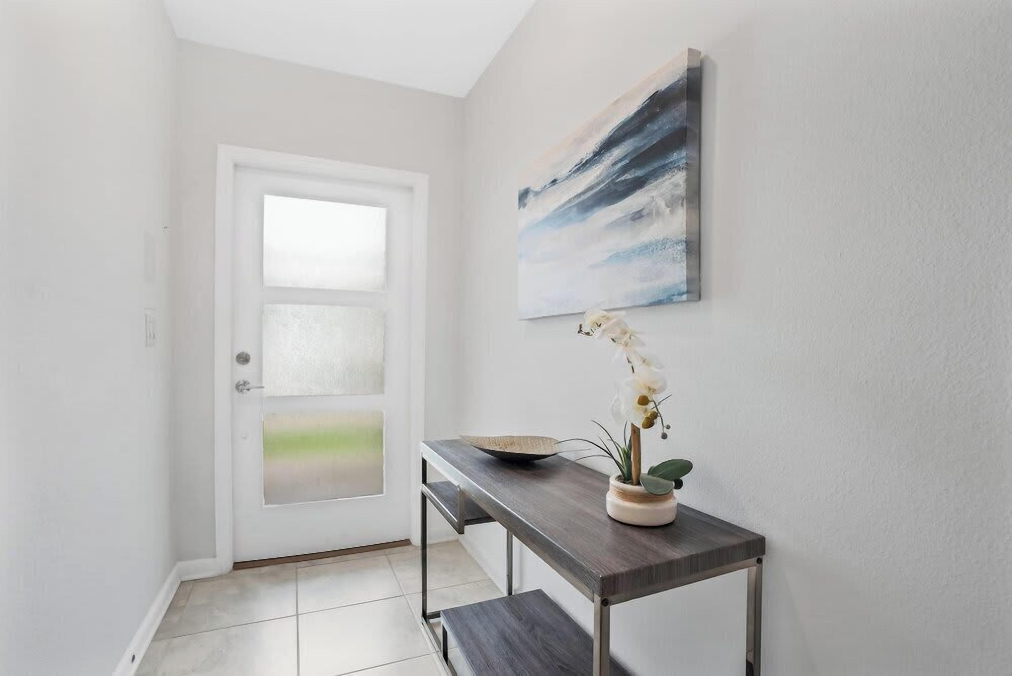 A hallway with a white door, a dark wooden bench, a small table with a bowl and orchid, and a painting on the wall.