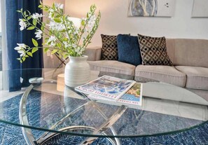A living room with a glass coffee table, a vase with white flowers, a beige sofa with patterned cushions, and a magazine on the table. - Family townhome near Disney w/ resort access (Davenport)