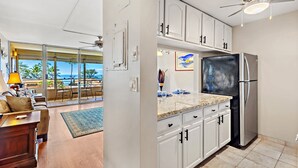 A modern kitchen with white cabinets, a stainless steel refrigerator, and a large window overlooking a scenic view. - In the Center of It all, walkable Kihei comfort (Kihei)