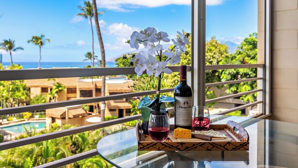A balcony with a glass table, a bottle of wine, a glass of red wine, a cheese and crackers tray, and a vase with white flowers. - In the Center of It all, walkable Kihei comfort (Kihei)