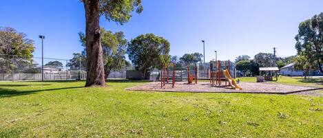 A playground with a slide and climbing structures, surrounded by a fenced area and trees.