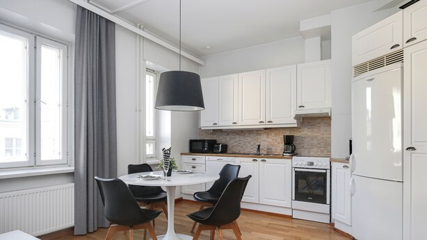 A modern kitchen with white cabinets, a dining table with chairs, a black pendant light, and a view of the city through large windows.