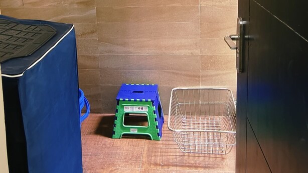 A small, narrow room with a tiled floor, a blue-covered table, a green stool, and a metal basket. - Sakthi Holiday's Inn (Chennai)