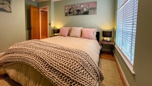 Quarto com uma cama grande, duas mesas de cabeceira com abajures, uma janela com persianas e uma porta.