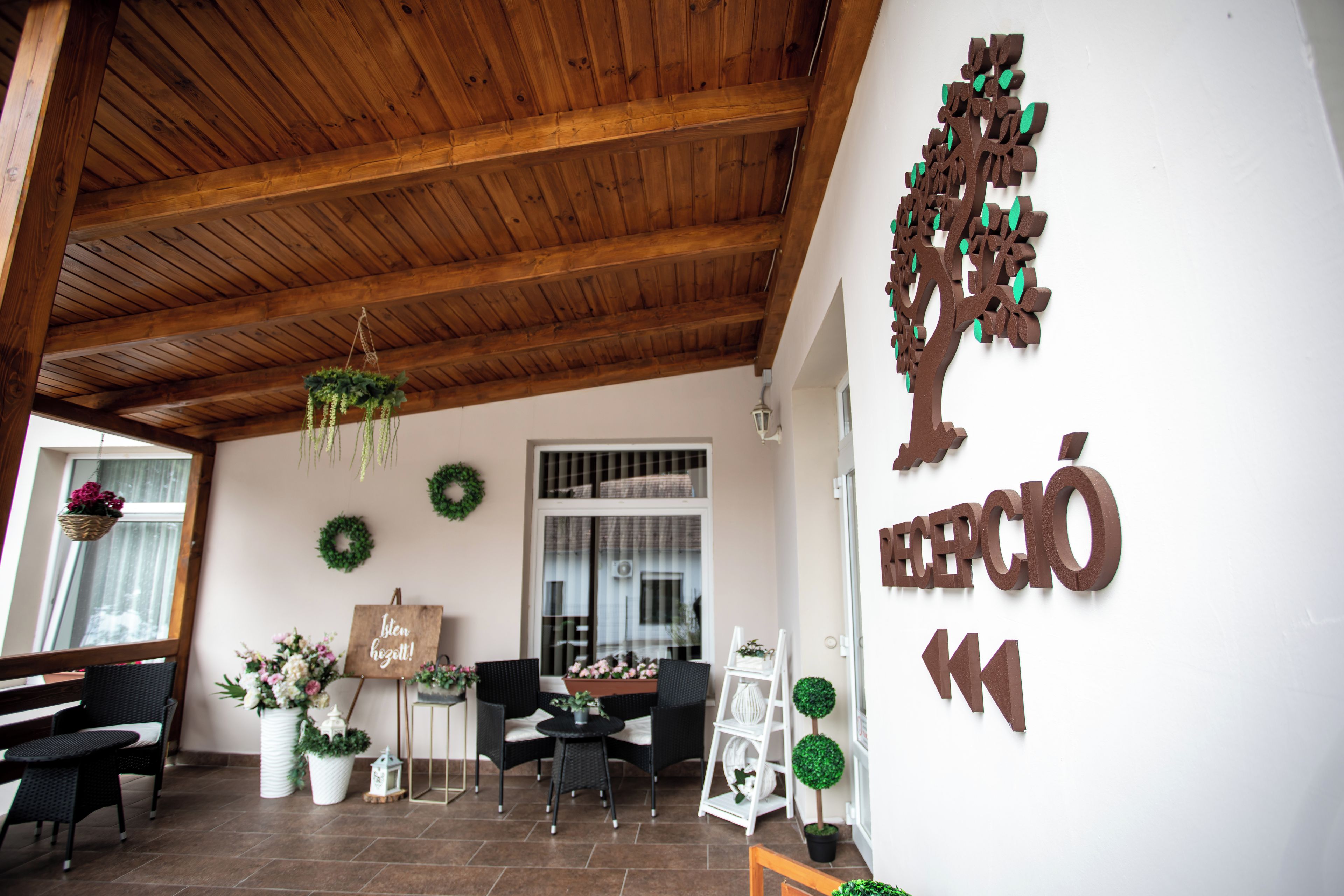 Un área de recepción con techo de madera, una mesa de recepción, sillas negras, una pared blanca con una gran decoración de árbol y un cartel que dice "RECEPCIÓN".