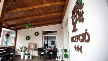 Un área de recepción con techo de madera, una mesa de recepción, sillas negras, una pared blanca con una gran decoración de árbol y un cartel que dice "RECEPCIÓN".