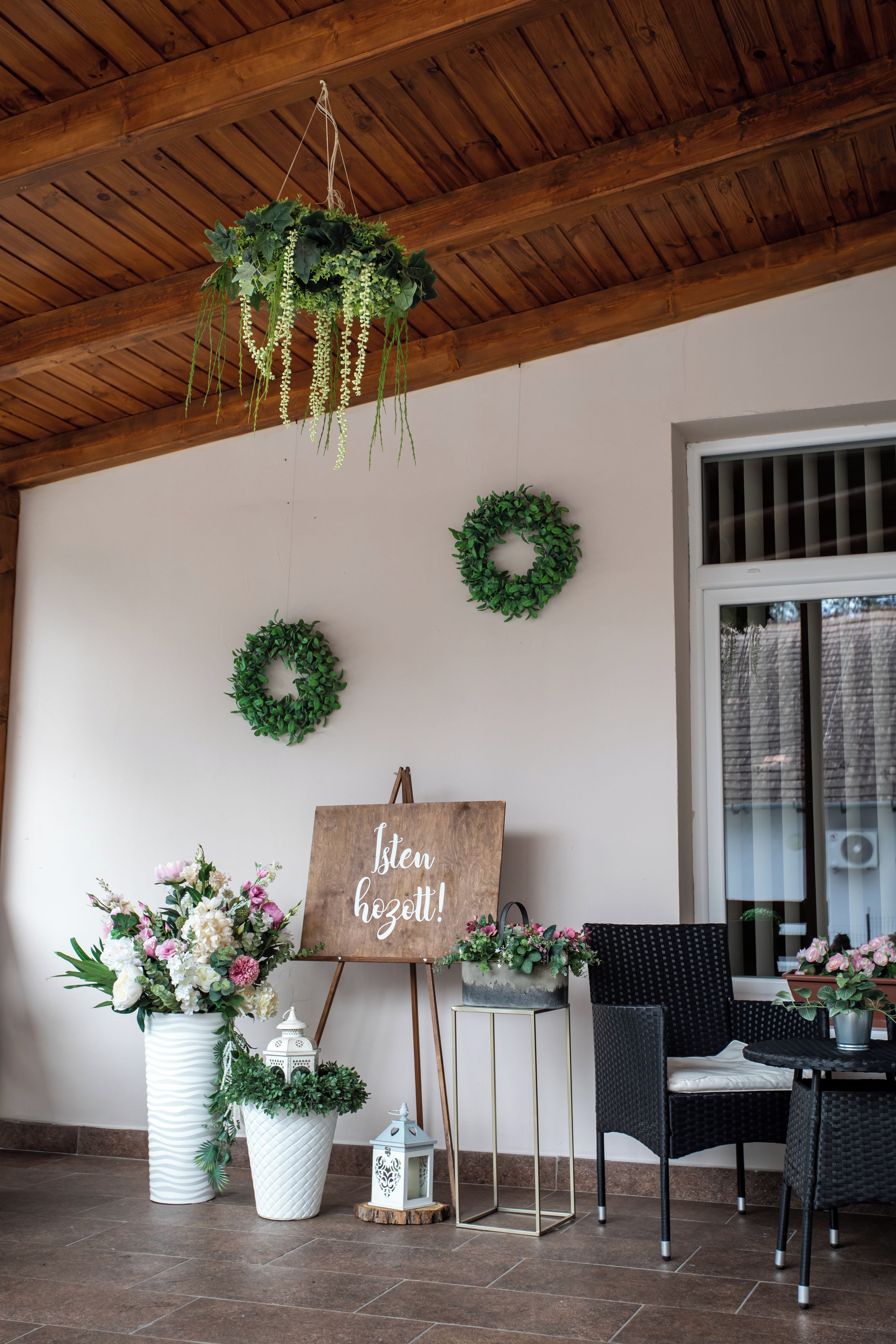 Una habitación con techo de madera, una pared blanca con dos coronas verdes, un caballete de madera con un letrero y una zona de estar con sillas y una mesa.