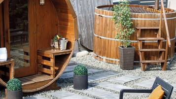 Una bañera de hidromasaje de madera, un barril de madera, asientos exteriores y una pequeña mesa con una planta y una botella.