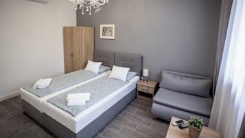A modern bedroom with a large bed, a grey sofa, a bedside table with a lamp, and a small potted plant.