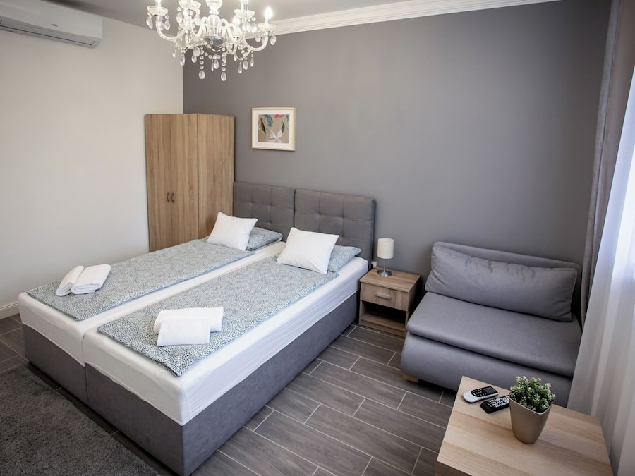 A modern bedroom with a large bed, a grey sofa, a bedside table with a lamp, and a small potted plant.