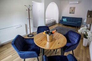 A modern living room with a round wooden table, four blue chairs, a grey sofa, a coat rack, and a bed in the background. - Árnyas Panzió Gödöllő (Gödöllő)