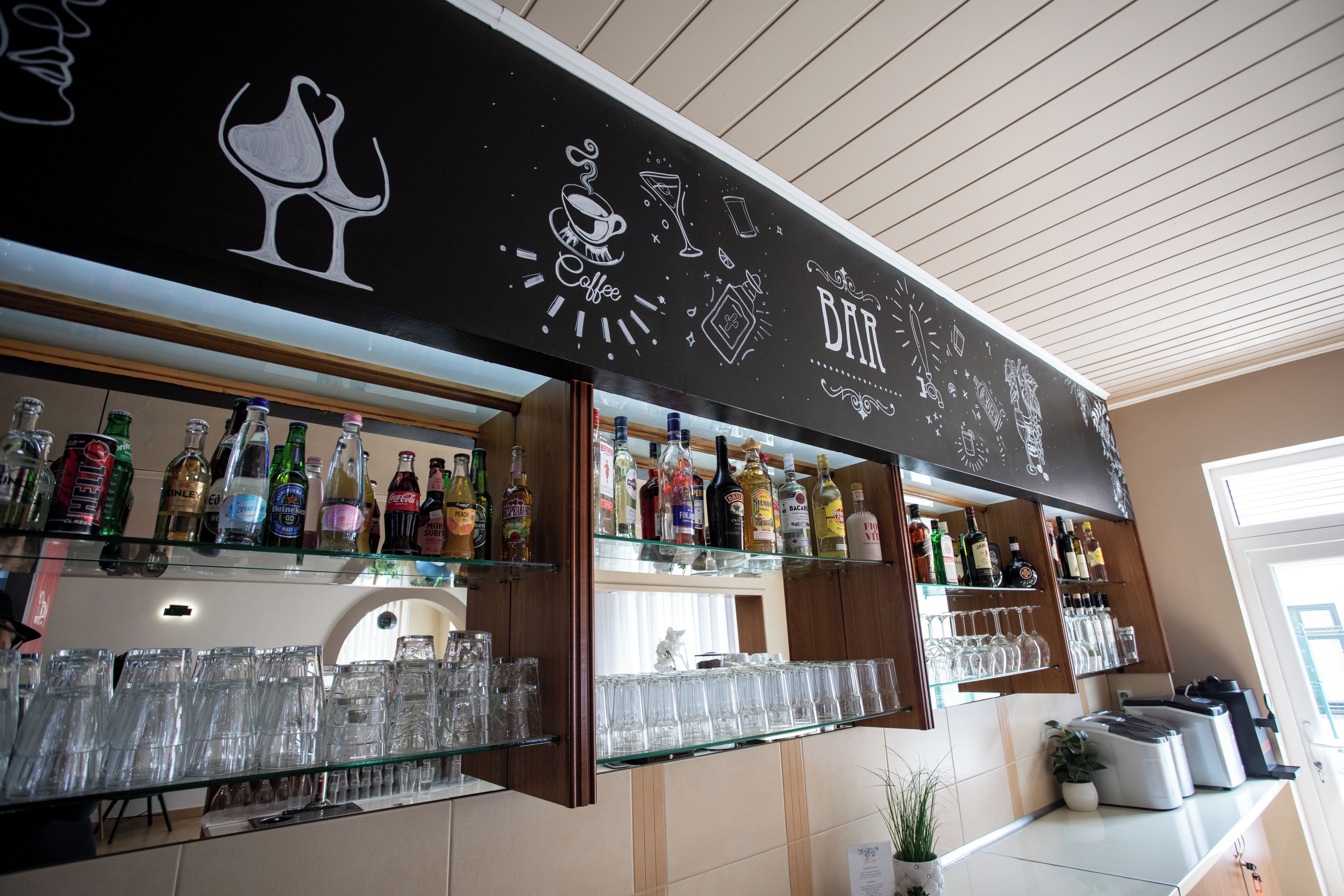 Una zona de bar con estantes llenos de botellas de licores y vasos, una pizarra con dibujos relacionados con la coctelería, y una planta en maceta sobre el mostrador.