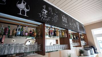 Una zona de bar con estantes llenos de botellas de licores y vasos, una pizarra con dibujos relacionados con la coctelería, y una planta en maceta sobre el mostrador.