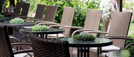 An outdoor seating area with wicker chairs and round tables, some with greenery on them.