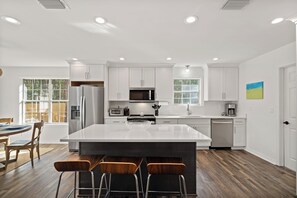 A modern kitchen with white cabinetry, a central island with stools, a refrigerator, microwave, and dishwasher. - Newly remolded home in the center of Dauphin Island (Dauphin Island)