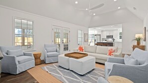 Un salon spacieux avec une grande fenêtre, un ventilateur de plafond et une cuisine à l’arrière-plan.