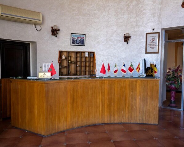 Una recepción con mostrador de madera, un cuadro enmarcado encima y un equipo de aire acondicionado instalado en la pared. Banderas decoran la pared detrás del mostrador.