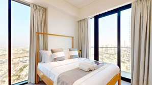 A modern bedroom with a large bed, a wooden headboard, and a view of a cityscape through large windows. - Two Bedroom: Modern and Chic with Balcony (Jumeirah Village Triangle)