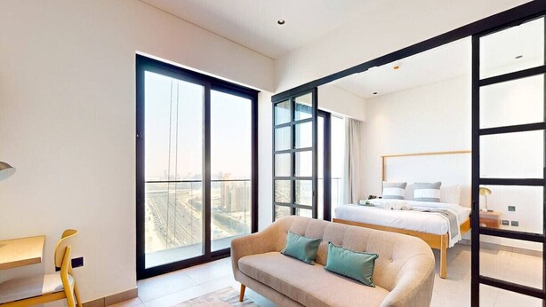 A modern hotel room with a large bed, a sofa, a desk, and a balcony with a city view. - Two Bedroom: Modern and Chic with Balcony (Jumeirah Village Triangle)