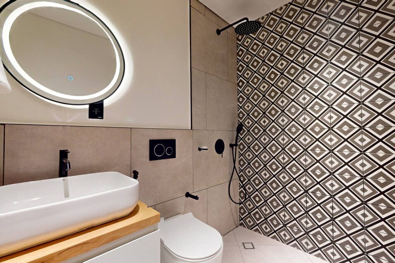 A modern bathroom with a white sink, a mirror with a circular light, a shower with patterned tiles, and a toilet.