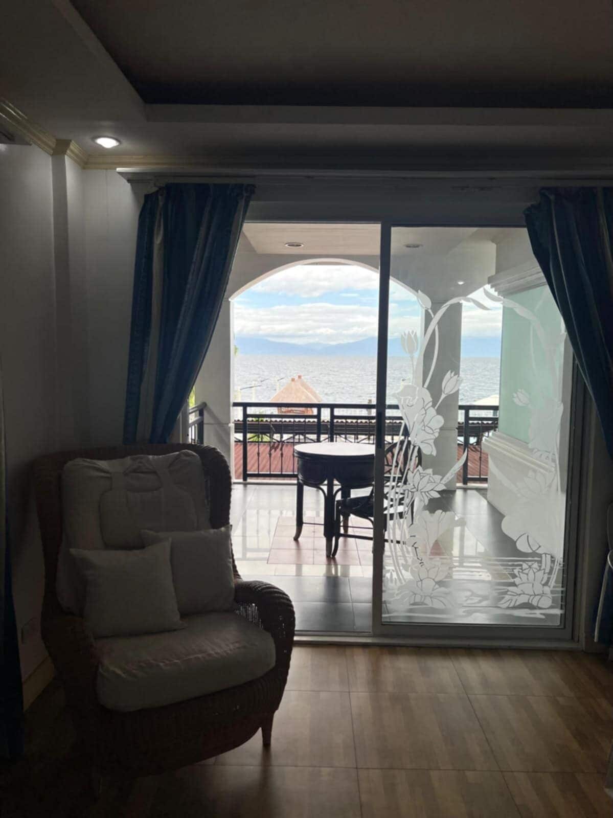 Een kamer met uitzicht op zee, een balkon met een tafel en stoelen, en een groot raam met gordijnen.