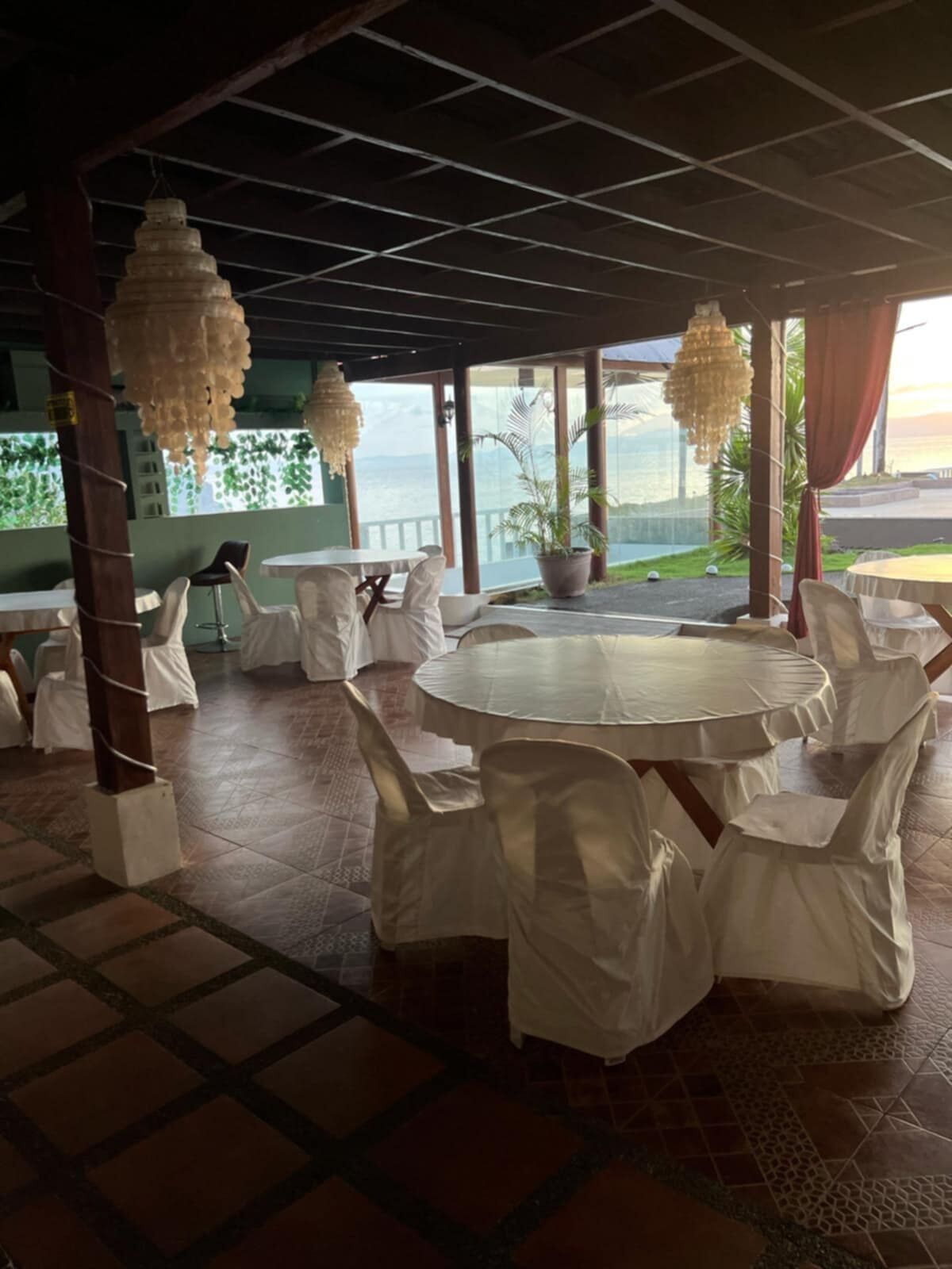 A covered outdoor dining area with round tables and chairs, large windows, and a view of the sea.