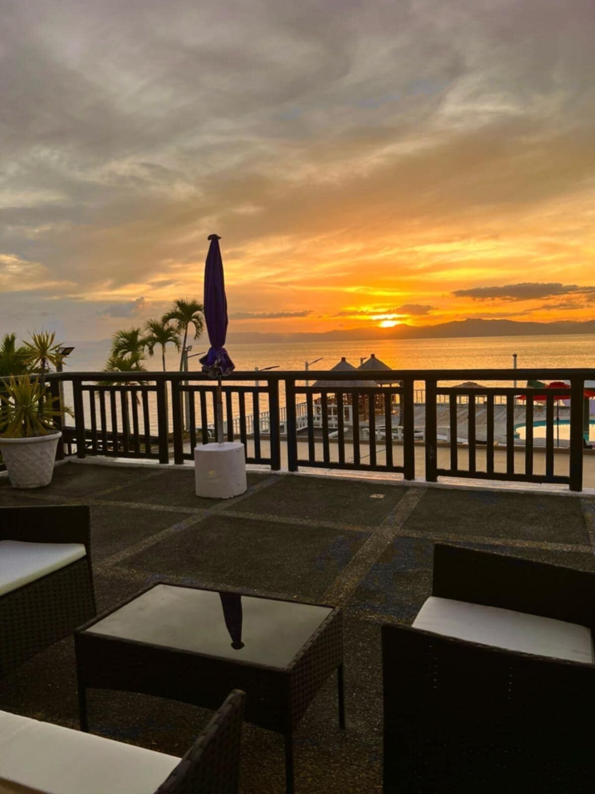 Een terras met zitplaatsen buiten, een uitzicht op de ondergaande zon boven de zee, en een hek.