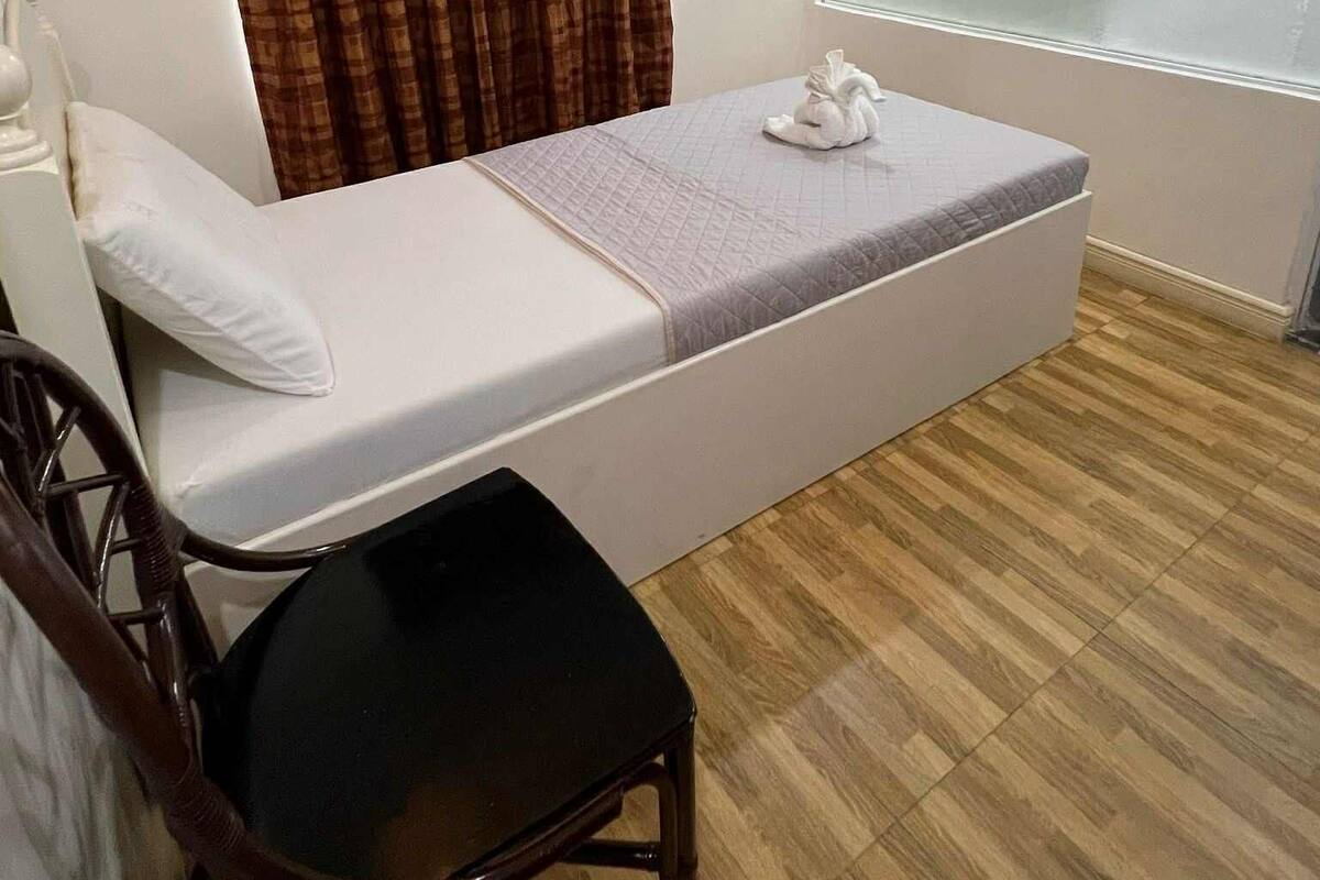 Ein Zimmer mit einem Einzelbett, Holzboden, einem weißen Bett, einem braunen Stuhl, einem Fenster mit Jalousie und einem Vorhang.