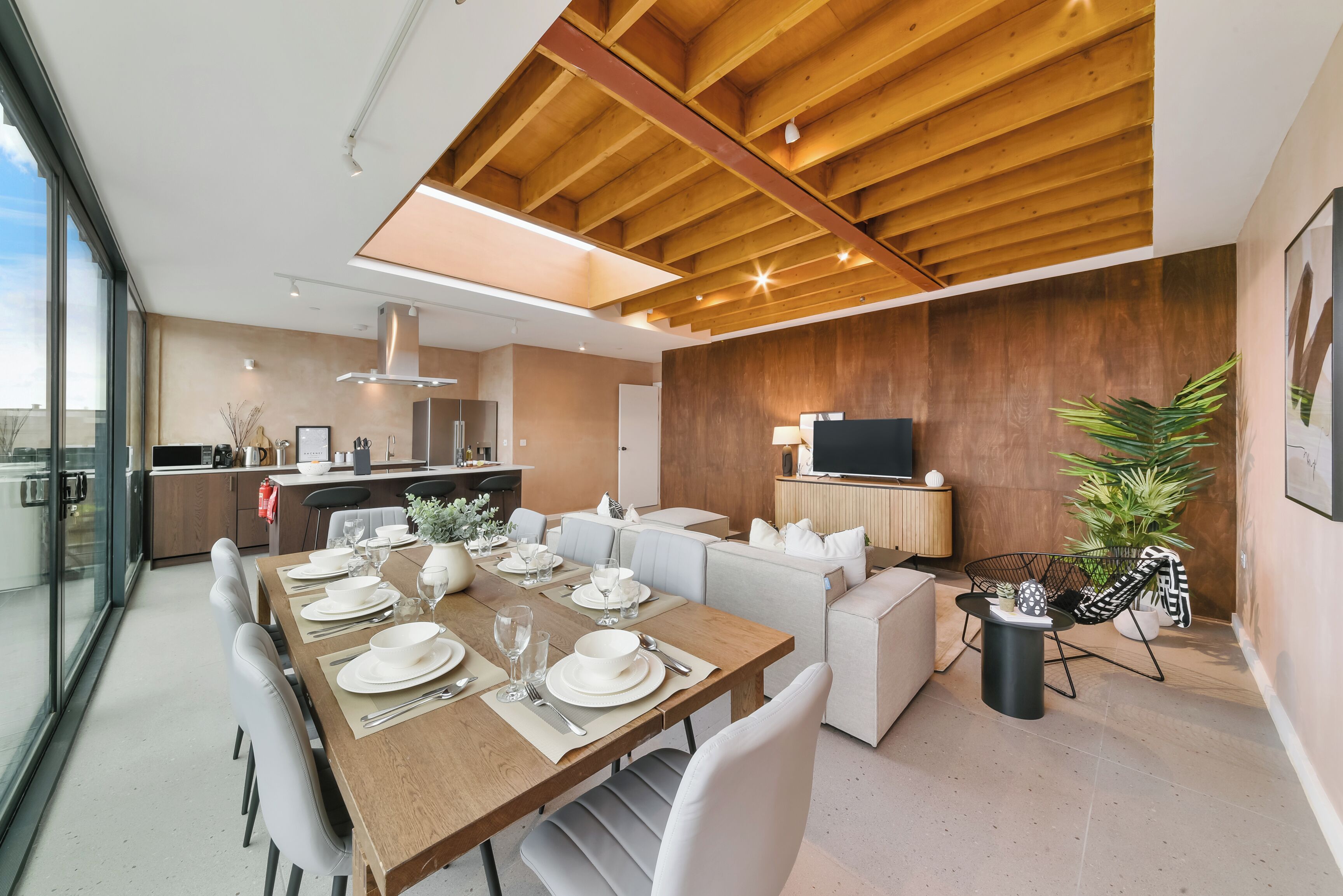 A modern dining area with a wooden table set for six, a sofa, and a TV in the background.