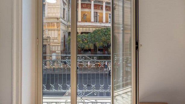 Une vue à travers une porte vitrée ornée d’une rambarde en métal, un bâtiment classique avec des colonnes et un balcon, et une rue animée de piétons et bordée de verdure.