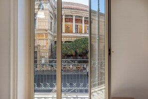 A view through a glass door with a metal railing, a classical building with columns and a balcony, and a street with pedestrians and greenery.