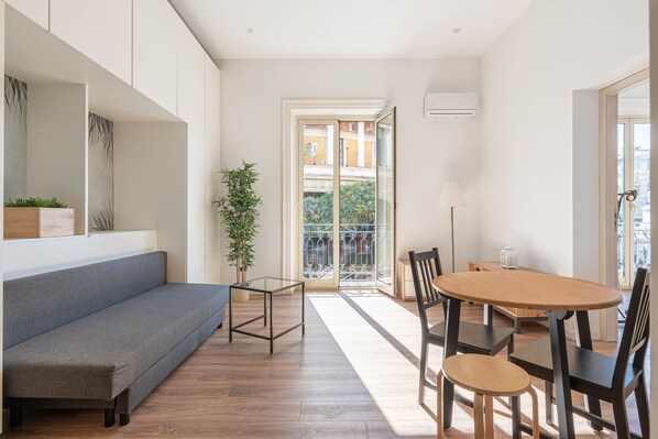 A modern living room with a grey sofa, a wooden dining table with chairs, a glass coffee table, and a potted plant.