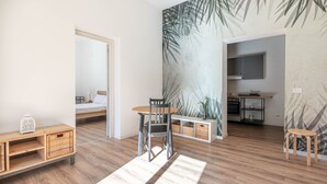 Une chambre moderne avec un plancher en bois, un coin repas comprenant une table et des chaises, un coin cuisine et un lit doté d’une tête de lit blanche.