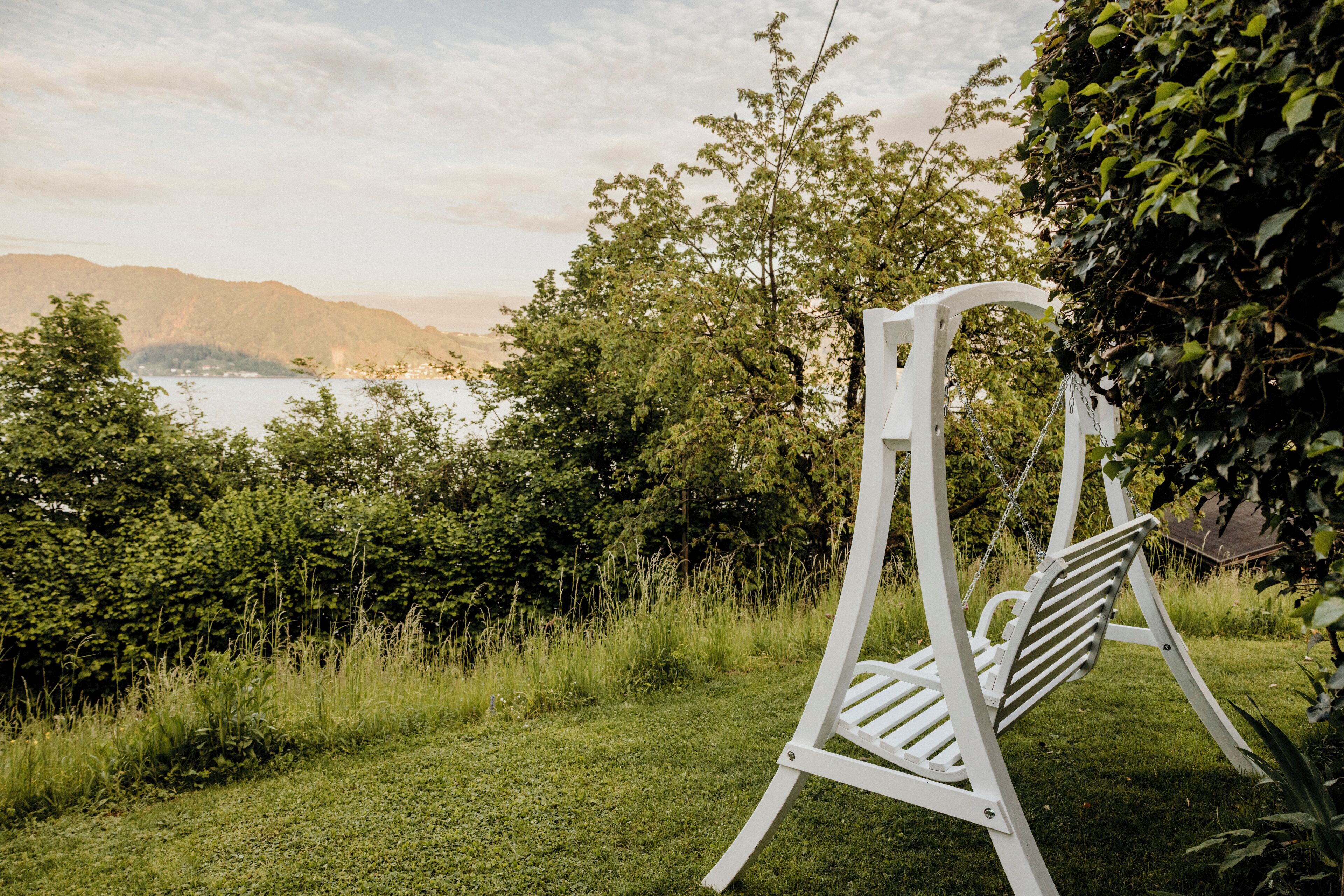 A white swing chair in a lush garden with a view of a lake and mountains in the background.
