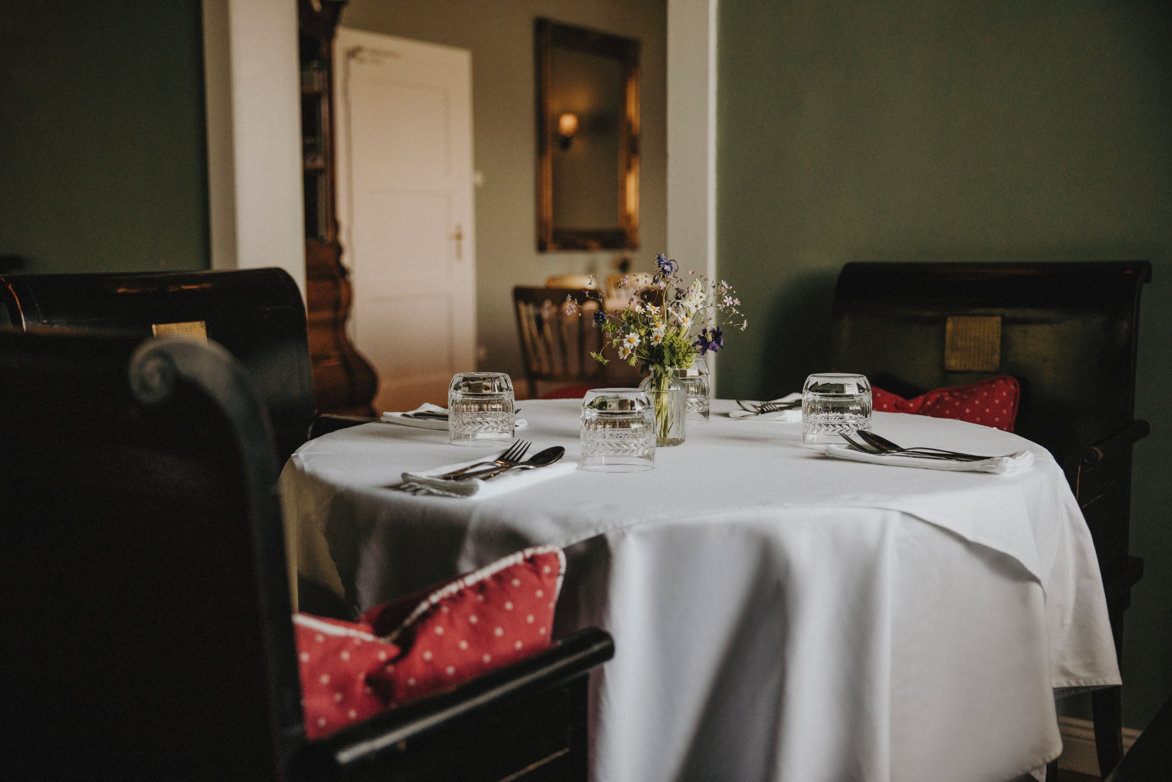 Une table à manger dressée avec de la verrerie et des couverts, un vase rempli de fleurs et une chaise agrémentée d’un coussin rouge.