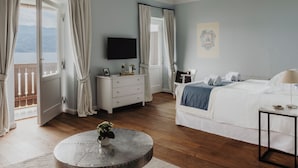 Une chambre à coucher comprenant un lit, une télévision, une commode, une table ronde avec un vase, et donnant sur l’extérieur.