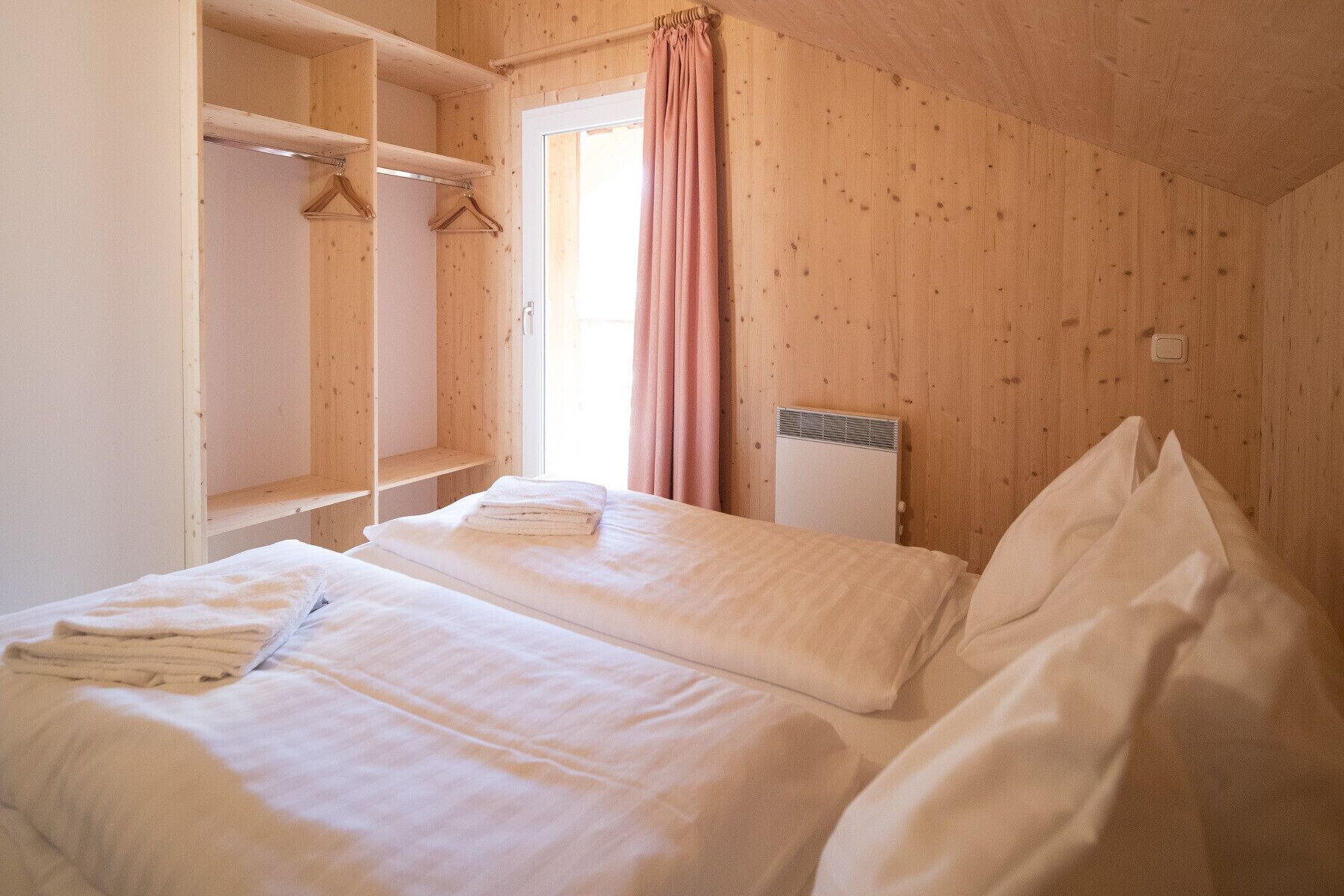 A bedroom with two beds, wooden walls, a window with pink curtains, and a radiator.