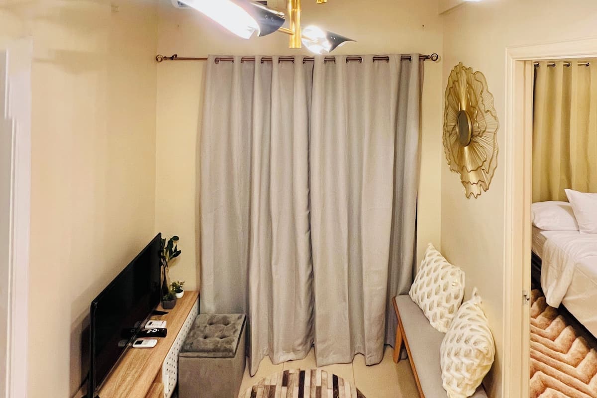A compact bedroom with a bed, a seating area, a TV, and a small table with decorative items.