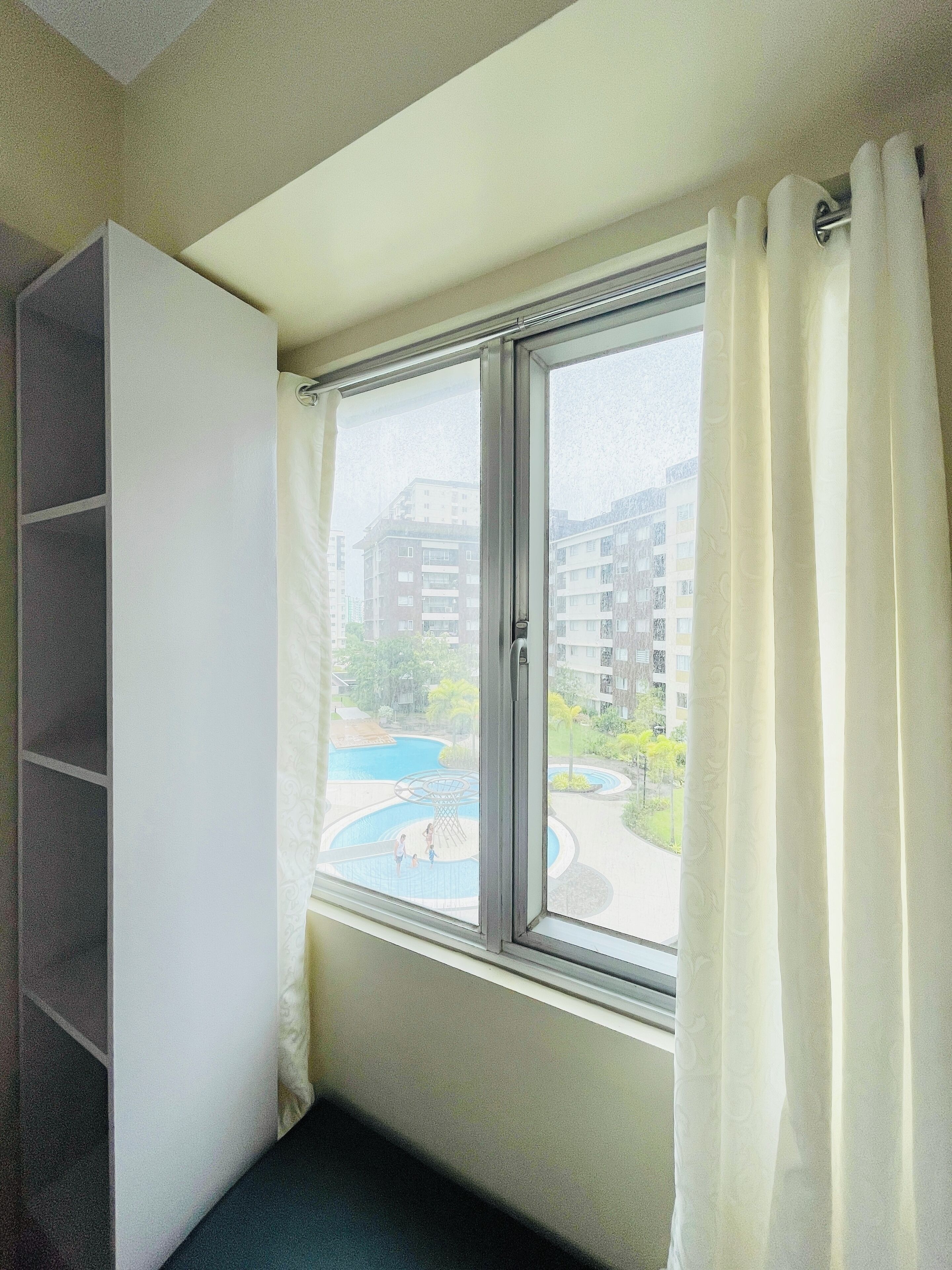 Una habitación con ventana, cortina blanca y vista a una piscina y edificios.