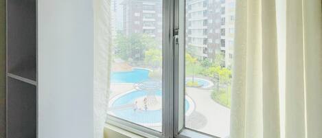 Una habitación con ventana, cortina blanca y vista a una piscina y edificios.